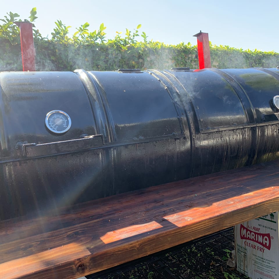 Large black cylindrical barbecue smoker with temperature gauges sits on a wooden stand outdoors beside Marina clear fry bags.