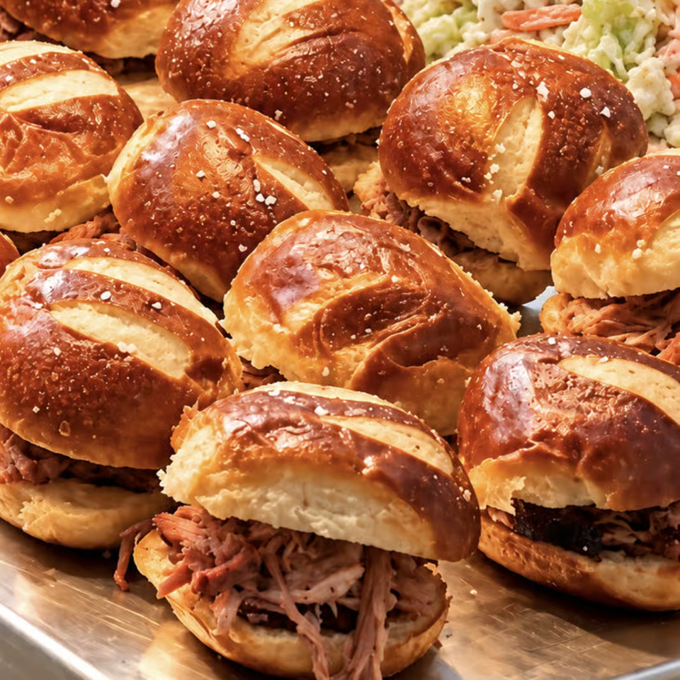 Tray of pretzel-bun sandwiches filled with pulled meat, served alongside creamy coleslaw.