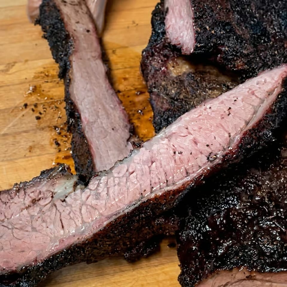 Sliced smoked beef brisket with a dark pepper crust rests on a wooden cutting board.